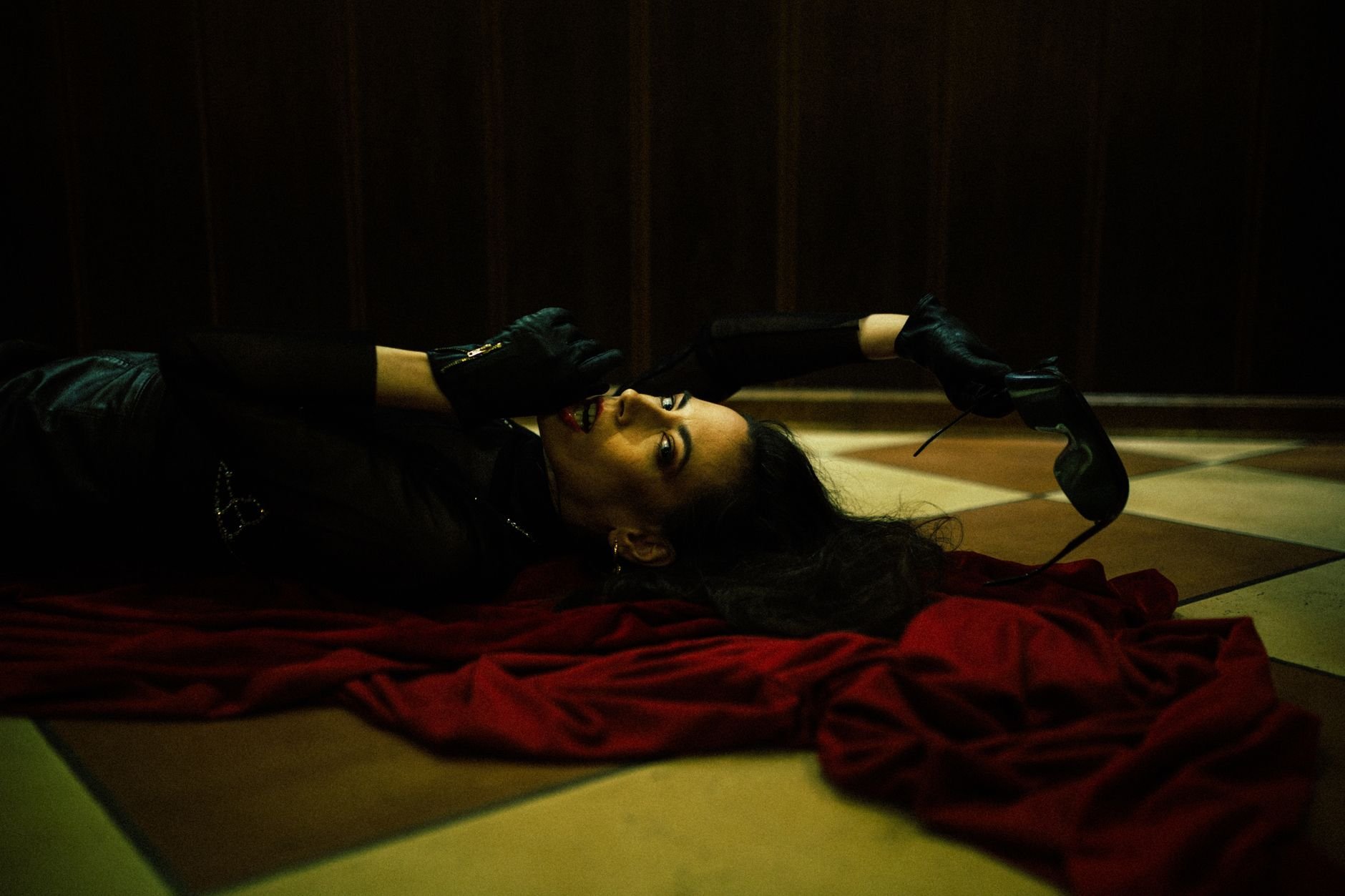 Fashion model in dark attire poses elegantly on red fabric indoors, creating a dramatic and artistic scene.